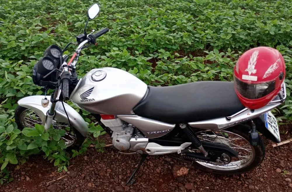 Motocicleta envolvida no acidente parada ao lado de uma lavoura de soja, com o capacete do condutor sobre o assento.